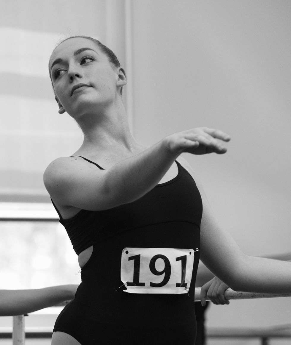 A young woman in a master class. She wears a black leotard with the number 191 pinned to her front. She is at the barre, with her outside arm extended in front of her and her head slightly angled out. We only see the top half of her body.