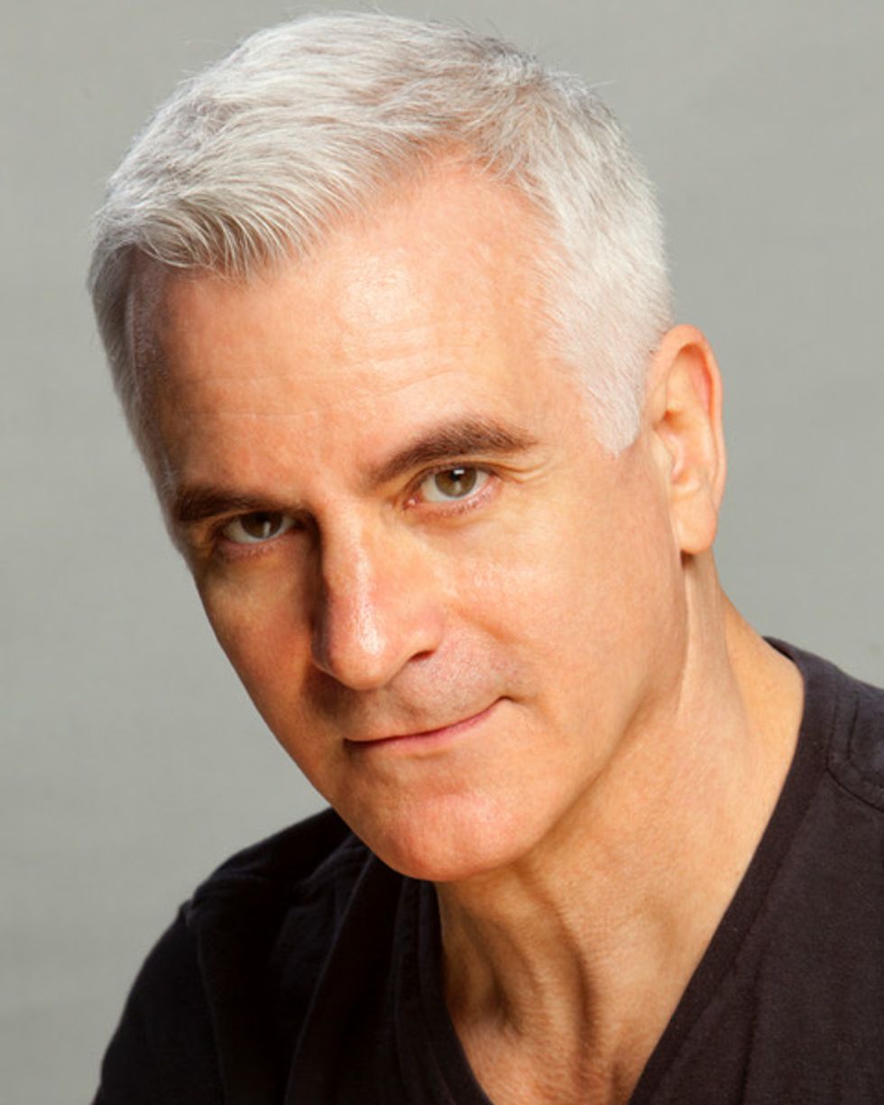 A man with white hair and a black V-neck T-shirt is shown from the chest up and looks directly at the camera.