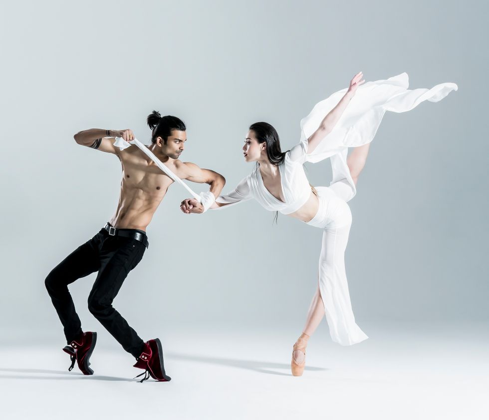 A male dancer (shirtless in black pants and sneakers) pliu00e9s while balancing on his toes. He is holding a female dancer's wrist. She is in a gauzy white top and pants and is doing a piquu00e9 attitude on pointe.