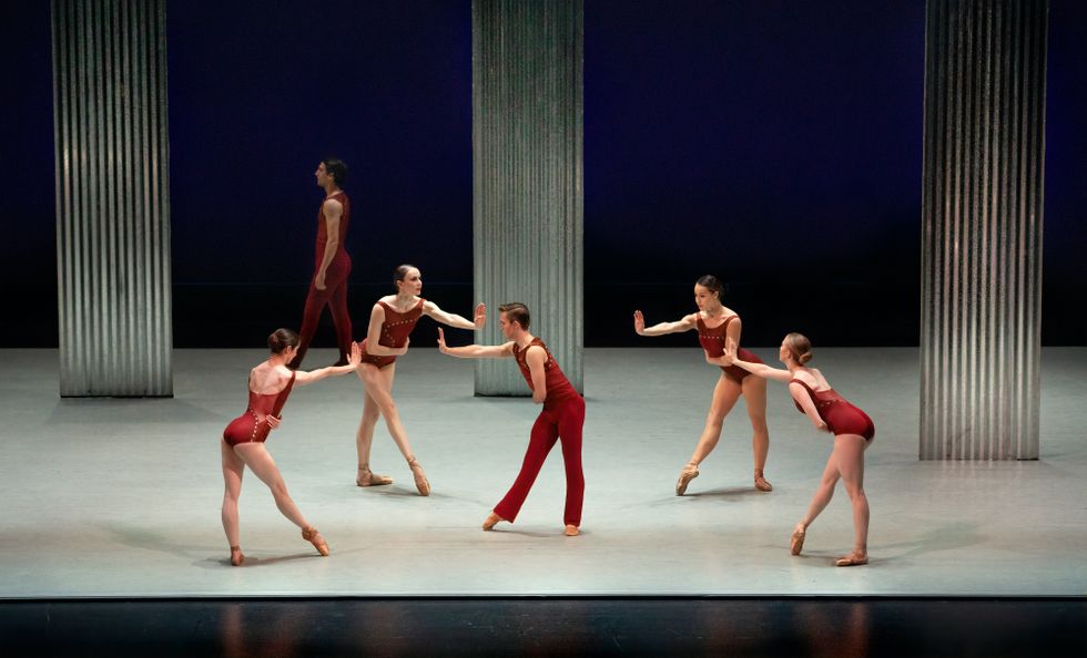 Four women, in deep red leotards and pointe shoes, encircle a male dancer also dressed in red. They gesture toward him with their right leg in tendu devant and right arm reaching forward with palm flexed upward.