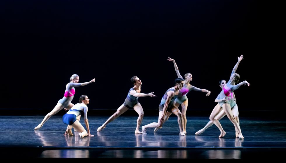 A group of six men and women in color-block leotards and short bodysuits gesture toward a woman at far stage left.