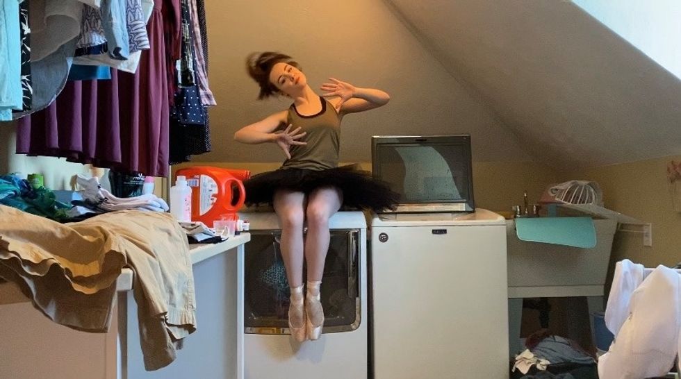 Wearing a black tutu, green tank top and pointe shoes, Kyra Laubacher sits on a washing machine and whips her head to her right, holding her arms in front of her.