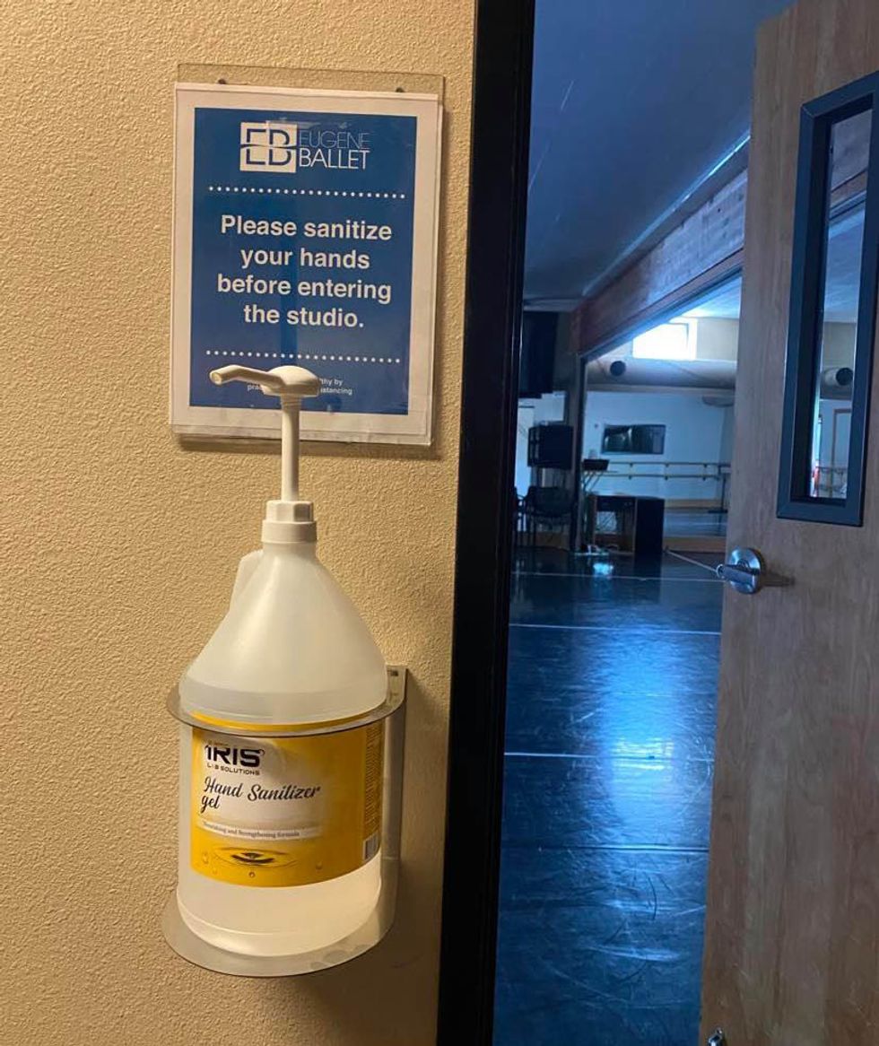 A large pump bottle of hand sanitizer sits in a dispenser attached to the wall, next to an open door that leads to a dance studio. Above the dispenser is a sign that says "Please sanitize your hands before entering the studio."