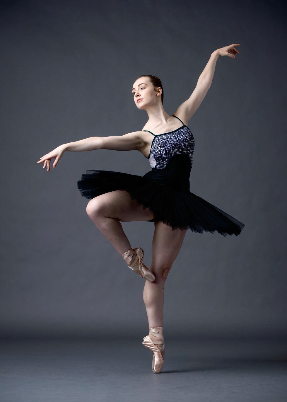 Wearing a black tutu, white and black patterned tutu and pointe shoes, Kyra Laubacher poses with her right leg in retiru00e9 and looks over her right arm.