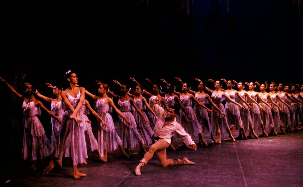 Lorraine Graves, wearing a purple dance dress and crown of flowers, stands on her left leg in tendu derriu00e9rre with her wrists crossed in front of her. Behind her stand a line of similarly dressed wilis in tendu, while Lowell Smith kneels in front of Graves.