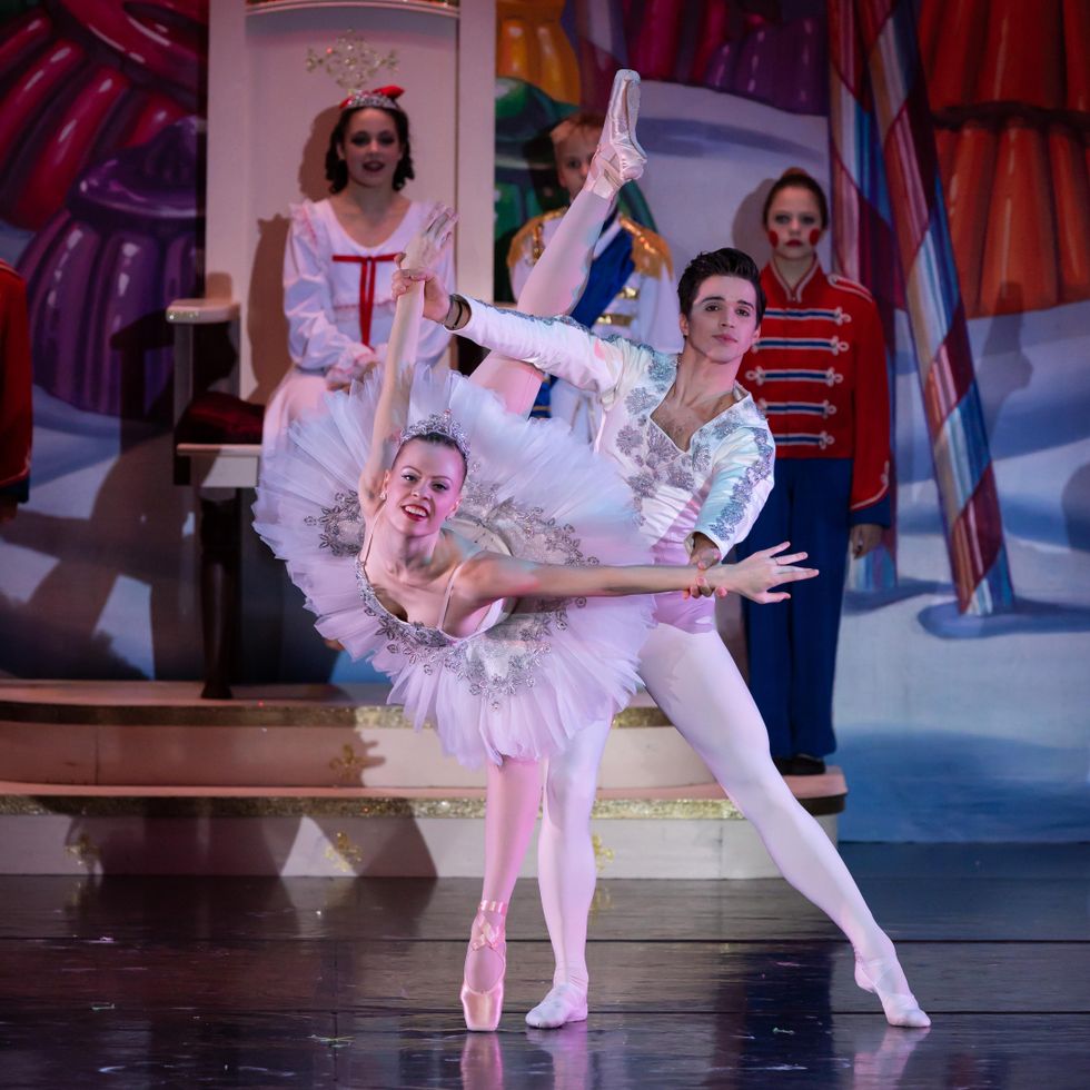 A ballerina in a sparkly white tutu, pink tights and pointe shoes performs a penchu00e9 arabesque on her left leg and presses her arms back. Her partner, wearing a matching jacket and tights, stands behind her and holds onto both of her wrists while three children in colorful costumes watch behind them onstage.