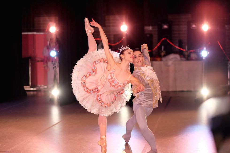 On a brightly lit stage, a ballerina in a pink tutu, tiara, pink tights and pointe shoes holds onto her partner's right hand and performs a penchu00e9 in second arabesque on her left leg. He wears a gray jacket with a white ascot tie and gray tights and lunges deeply on his right leg while holding his left arm out to the side.