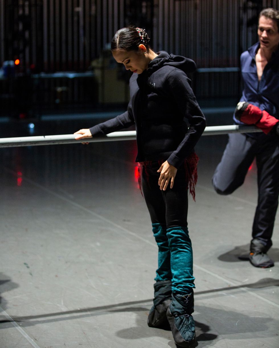 Pantastico, dressed in warm up clothes and booties, stands at the barre in first position relevu00e9, looking down at her feet.