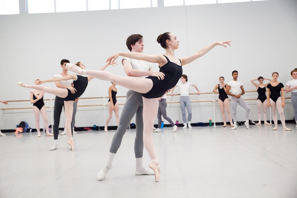 A partnering class, with two couples in the center of the studio and the rest of the students watching in the back. The young men support the young women in a first arabesque.