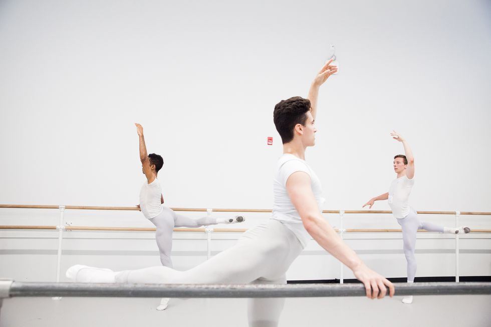 A group of young men taking class. They wear grey tights and white shirts. They are in an attitude at the barre.