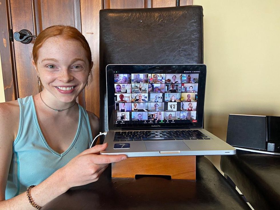 Zimmi Coker poses next to a laptop showing a ballet Zoom class and smiles brightly for the camera. She wears her red hair in a low bun and a mint green tank top.