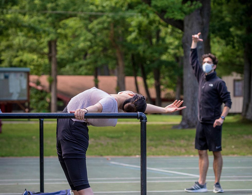 Outside on a tennis court, a female ballet student in a pink sweater and gray sweatshorts practices cambru00e9 back at a portable barre. Behind her in the the distance is her male instructor, wearing a face mask and workout clothes, who watches her and holds his right arm up to mimic hers.