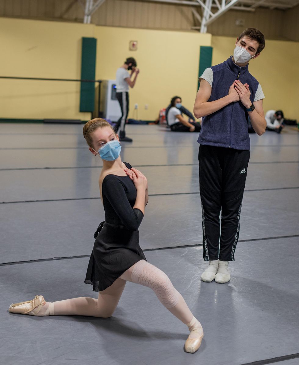 In a large dance studio, a female dance student in practice clothes and a blue face mask kneels on her left leg and holds her hand to her heart. Next to her on the right, a young male student in track pants, a blue mask and a fleece vest stands in parallel and holds his hands to his heart. They both look at their reflection in the mirror (not shown).