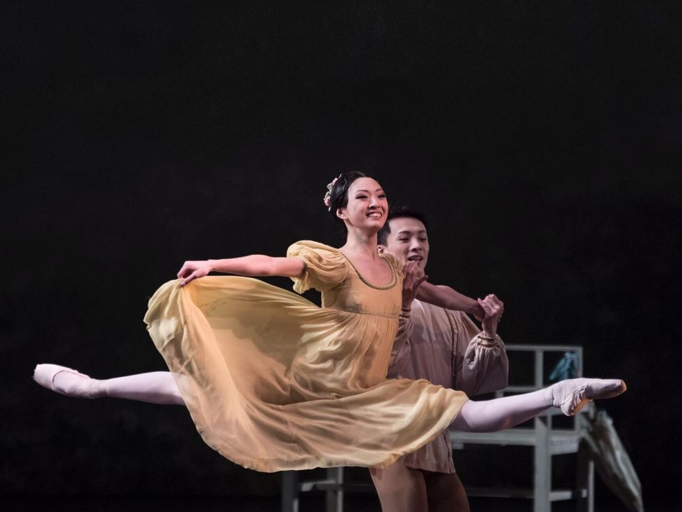 Patricia Zhou, wearing a big smile, a beige empire-waist dress and a floral headpiece, does a partnered grand jetu00e9 to the left, holding onto her male partner's left hand with her own left hand. She holds her skirt out to the side with her right hand.