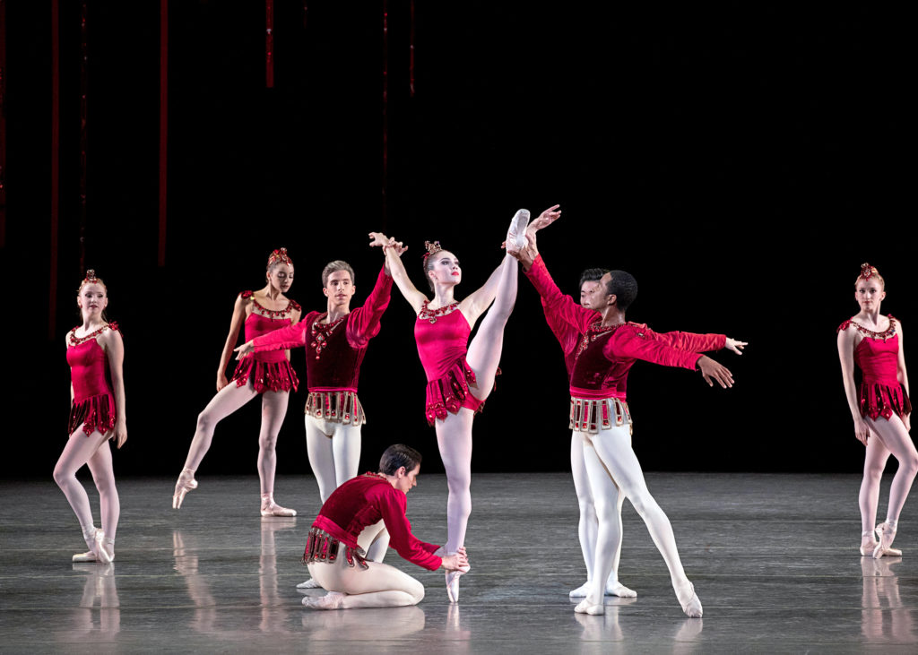 Mira Nadon stands on her right leg on pointe and does a développé ecarté with her left. A group of male dancers cluster around her, with one man on his knees holding her ankle and three others standing and holding her wrists and left foot. Three female dances stand in the background onstage. All the women wear short, sparkly red dance dresses while the men wear red jackets and white tights.