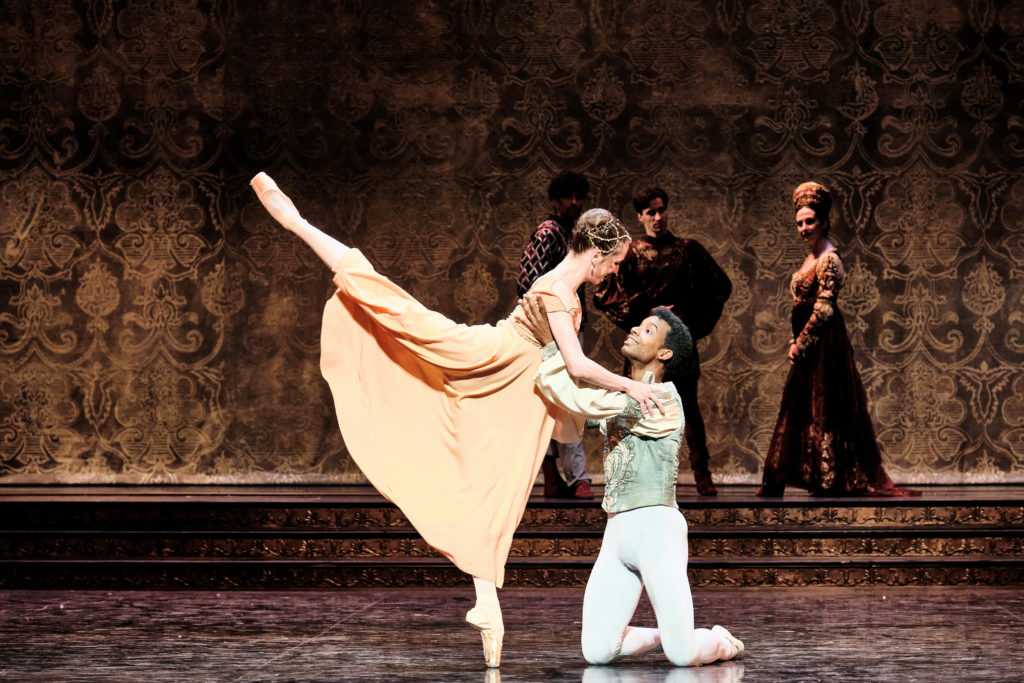 Gouneo, kneeling, holds Colasante as she balances en pointe in arabesque.