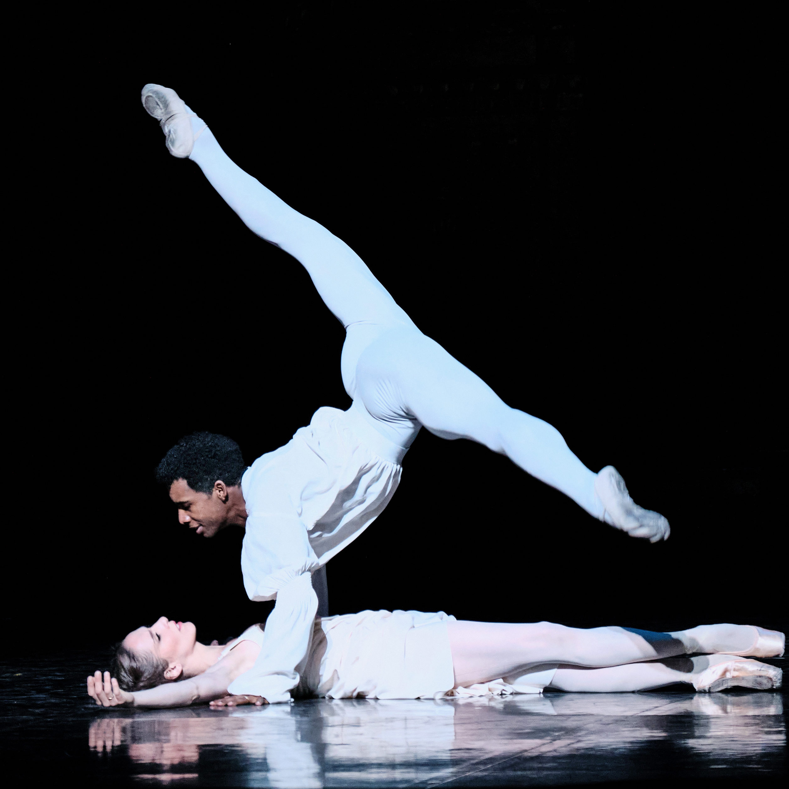 Osiel Gouneo and Valentine Colasante, in white costumes, perform onstage. Gouneo is in a split position balanced on his hands, looking at Colasante, who is lying on the stage beneath him.
