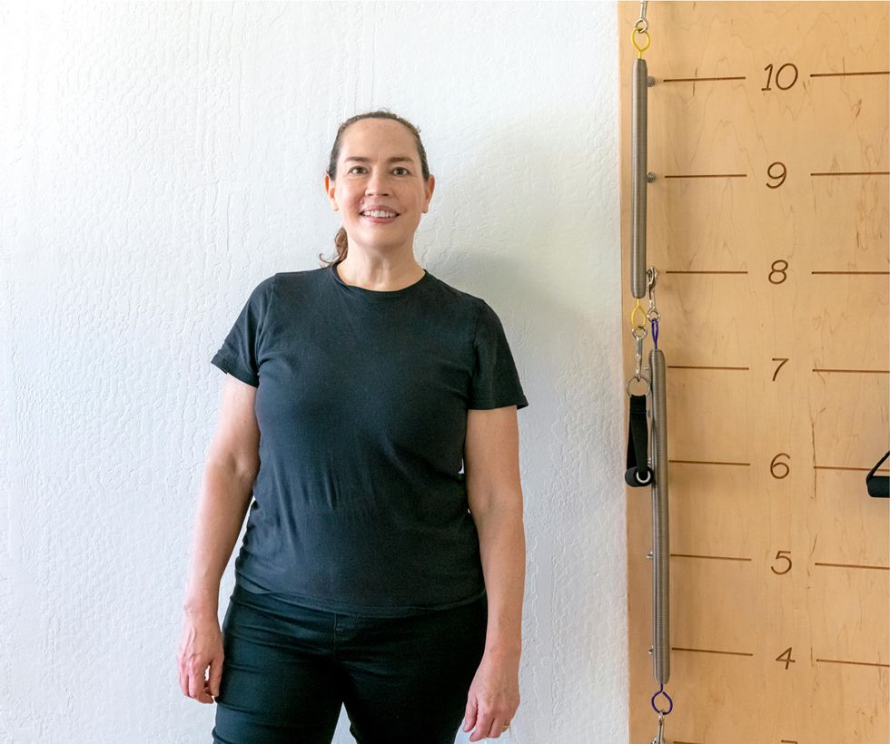 Kendall Alway, smiles, while standing in her physical therapy studio next to a wall equipped with resistance springs.