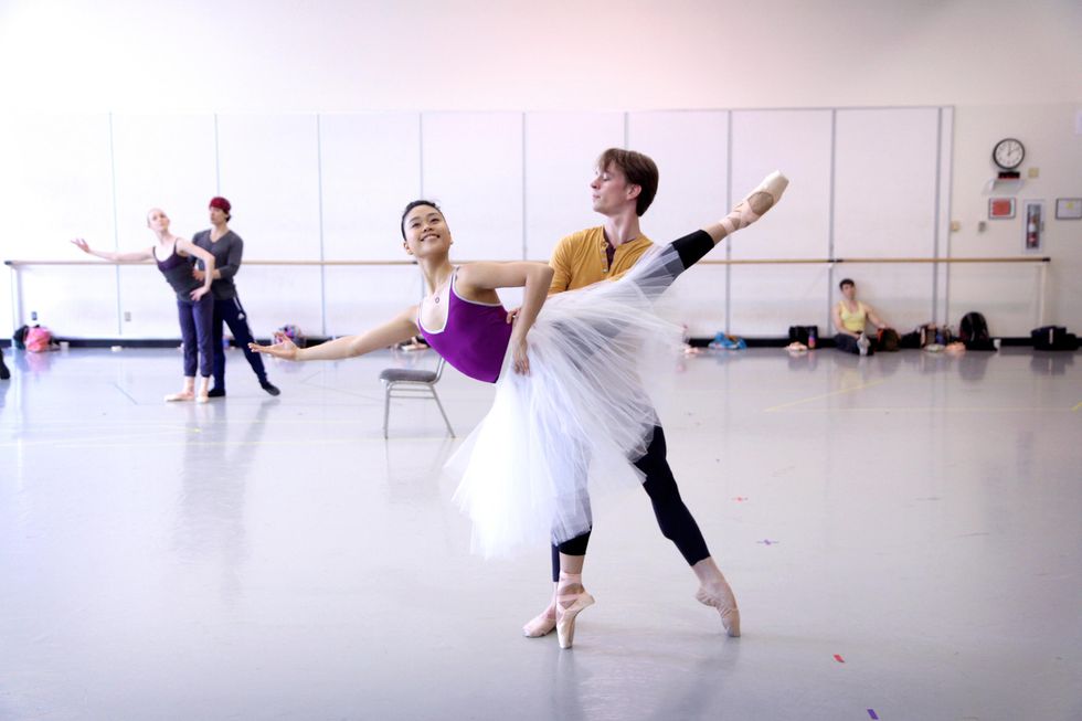 Angelica Generosa performs an arabessque elongu00e9 on pointe while her partner stands behind her holding her waist and with his left leg in tendu. She holds her left hand on her hip and extends her right arm out to the side with her palm up. Angelica wears a purple leotard, black tights and a white Romantic tutu while Kyle wears a yellow shirt, black tights and tan slippers.