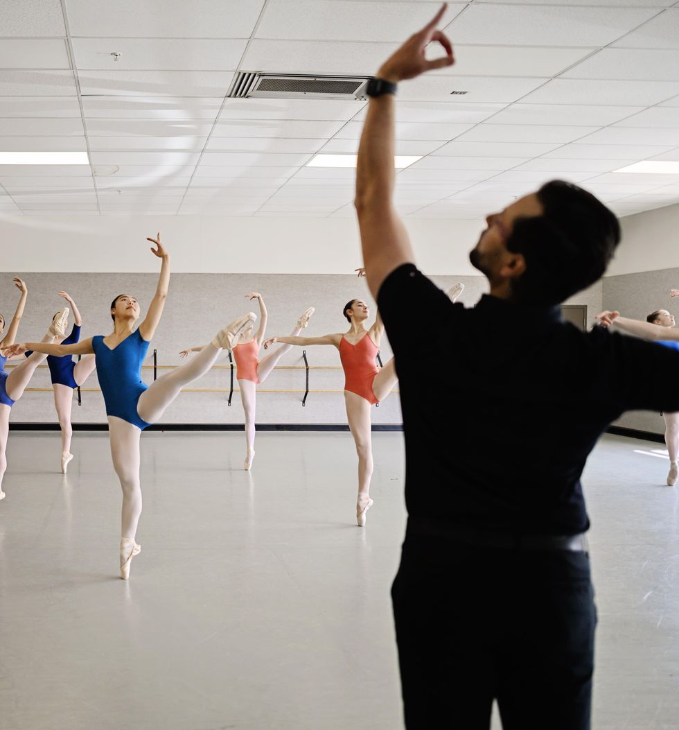 Simon Ballet, wearing dark clothing, is shown from behind demonstrating ecartu00e9 arms while in front of him, a class of teenage ballet students perform du00e9veloppu00e9 ecartu00e9 devant on pointe in a medium-size studio. The dancers, all girls, wear leotards, pink tights and pointe shoes.
