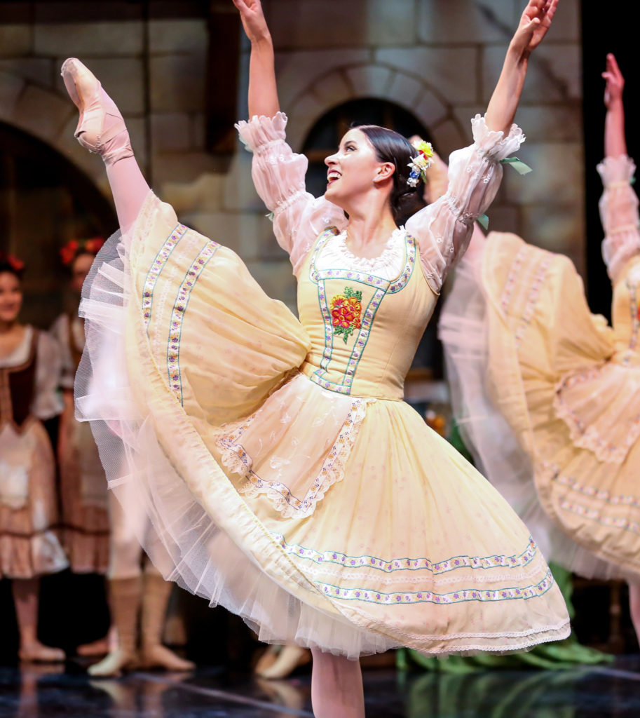 Wearing a yellow and white peasant dress, Alana Griffith does a second development with her right leg and raises her arms in a V-shape, looking up to her right hand.  She dances on a crowded stage at a city scene in a classical ballet.