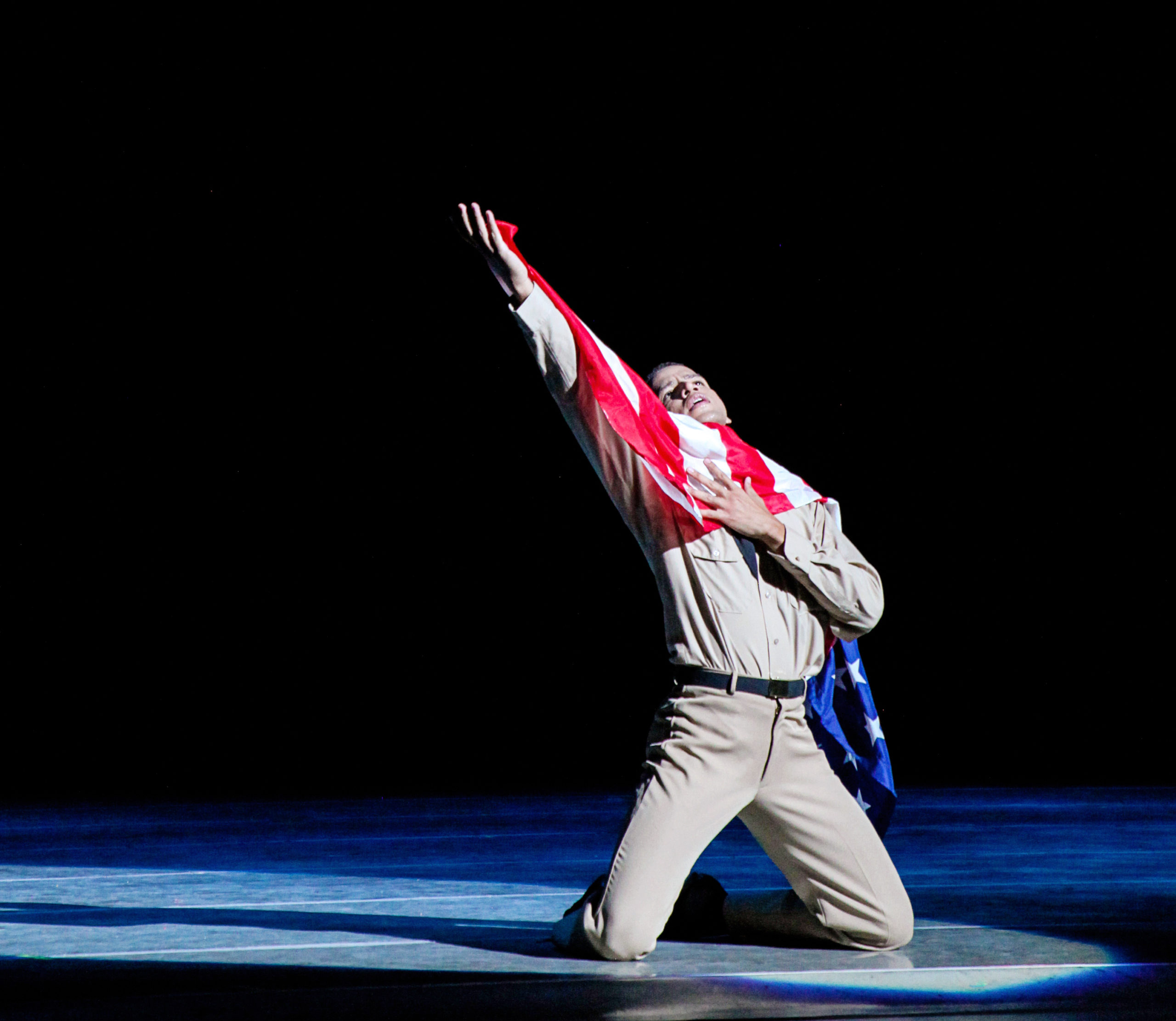 Wearing khaki pants and a a khaki button down, a spotlit Andres Castillo kneels on the ground onstage and reaches up with his right arm. He holds an American flag in his right hand, which drapes over his left shoulder and down his back, and looks up towards his hand.