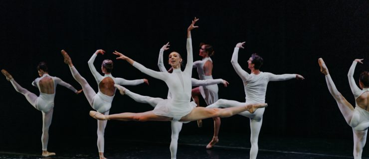 Ally Helman, wearing a white turtleneck leotard and skirt, performs a saut de chat center stage with a joyous smile. Behind her, a group if male and females dancers in white unitards perform develeoppé ecarté facing upstage.
