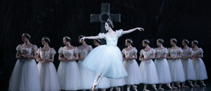 Sae Eun Park, wearing a white Romantic tutu and crown, performs a high assemblé in front of a diagonal line of female corps dancers. Behind the dancers onstage is a large cross.