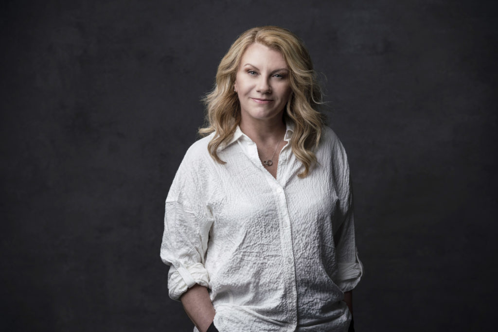 Hope Muir is shown from the waist up in front of a dark backdrop, smiling to the camera and with her hands in her pockets. She wears a textured, white button-down shirt and her shoulder-length blond hair down.