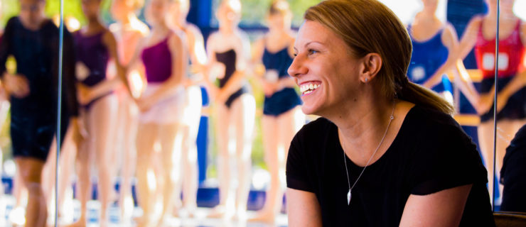 Hope Muir, in a black T-shirt and wearing her hair in a low ponytail, smiles widely as she looks towards company dancers in class. She is shown from the waist up, sitting down, and the dancers are shown in the reflection of floor-to-ceiling mirrors.
