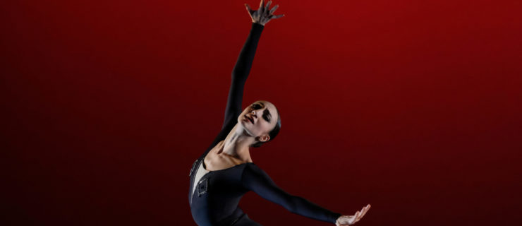 Katia Raj is shown from the thigh up in croisé, standing on her leg leg in plié and spreading her arms out wide with splayed fingers. She looks out into the audience dramatically, and wears a dark velvet leotard with rhinestone decor and dark tights.