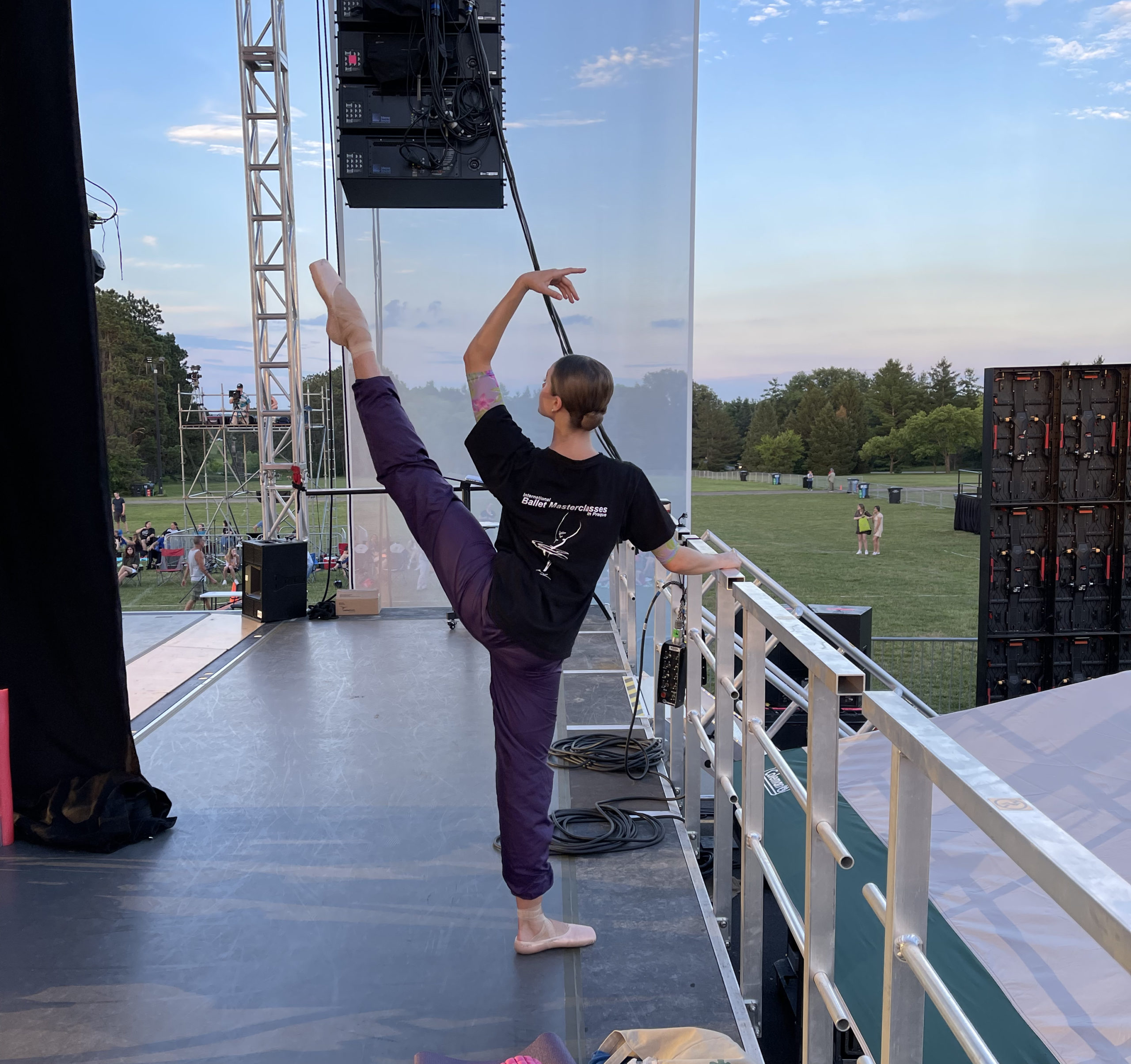 Abbey Marrison is shown from the back on an outdoor stage, holding on to a barre and doing a grand battement second with her left leg. She wears purple warm-up pants, a black T-shirt and pink pointe shoes.