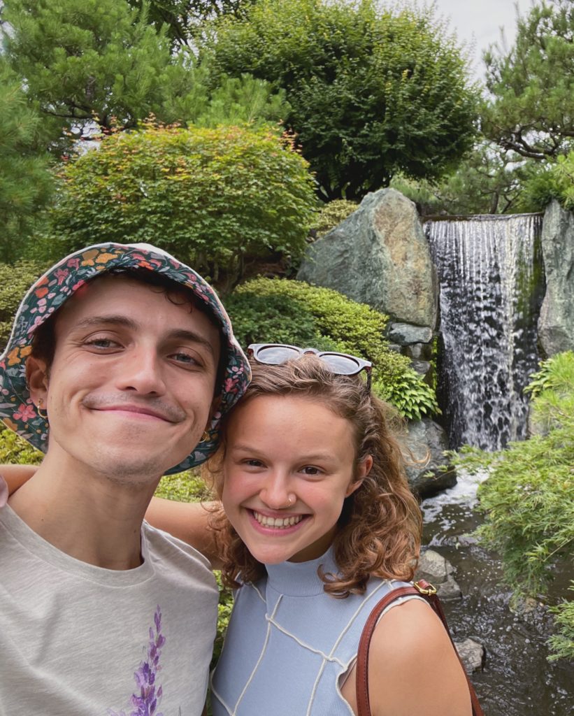 Jacob Clerico and Abbey Marrison pose for a selfie in front of a waterfall and trees. He wears a floral print bucket hat and white T-shirt, while Abbey wears a blue sleeveless turtleneck and a pair of sunglasses on her head.