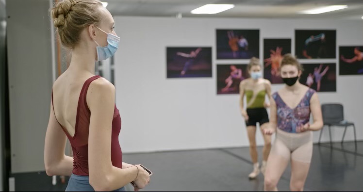 Ally Helman, in a burgundy leotard and face mask, stands at the front of a dance studio while two female dancers in the background mark through movement.