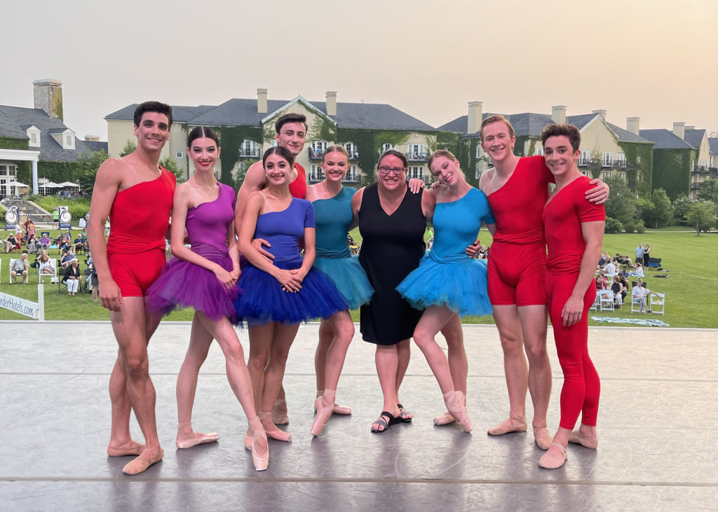 A group of eight male and female dancers and one female musician in a black dress pose with their arms around each other onstage. The men wear red costumes while the women wear blue, purple or green tutus. Behind them is a green lawn with white chairs and audience members.