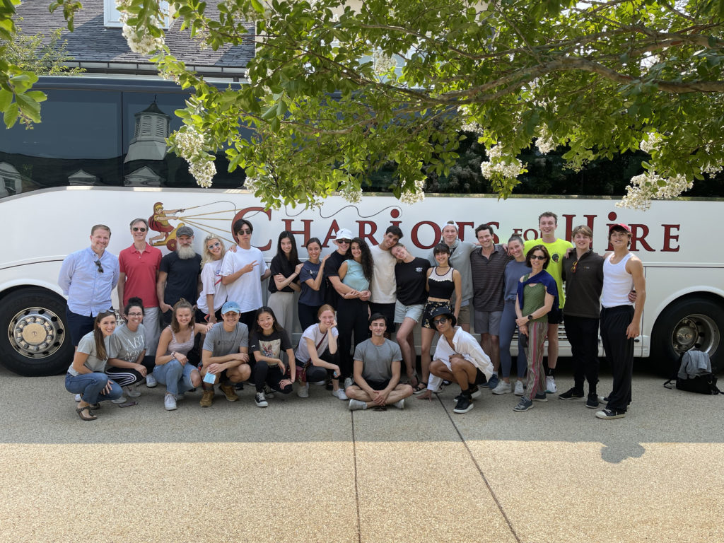 A group of 27 men and women group together for a group photo in front of a large charter bus.