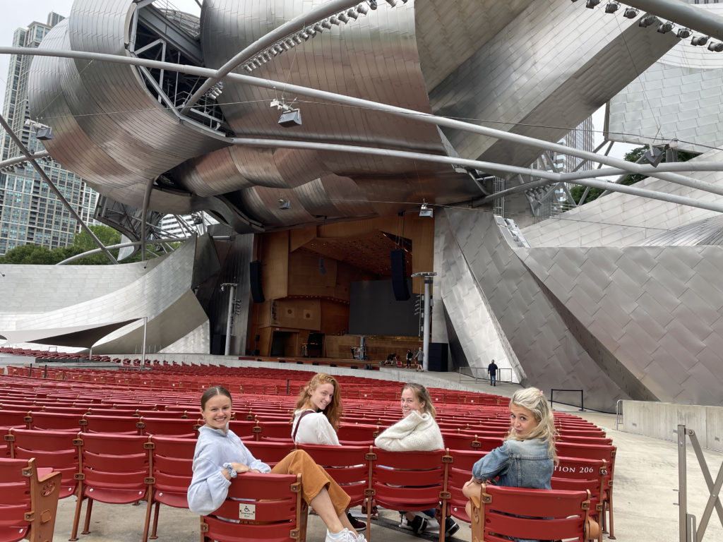 Four young women in street clothes sit in red chairs in front of a large, modern-looking stage.