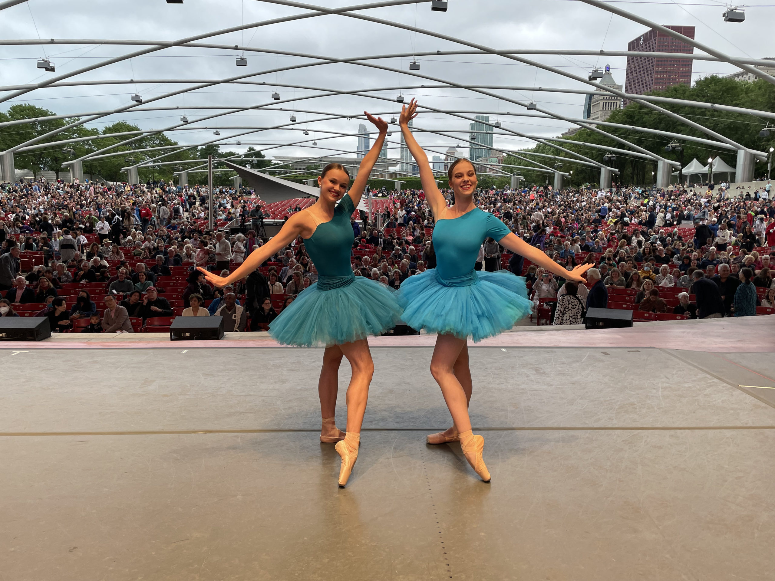 Two female dancers wearing turquoise, one-shouldered tutus, stand opposite each other and bevel their foot on pointe, extending their arms wide with flexed hands. They stand on an outdoor onstage, with a milling audience behind them.