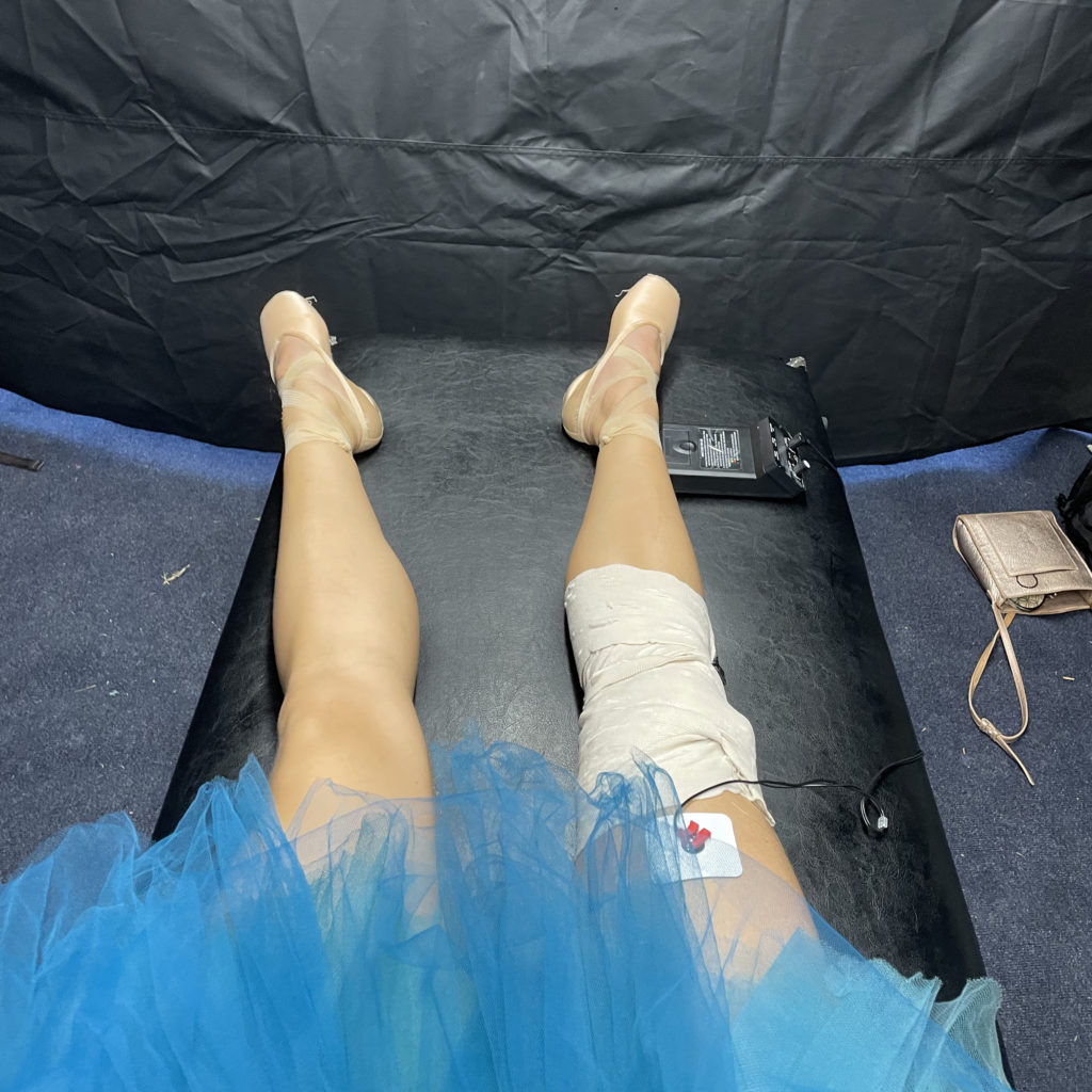 A female ballerina's legs are shown on a physical therapy table with a bandage wrapped around her right knee and an e-stim machine attached to her thigh. She has pink pointe shoes on and is wearing a turquoise tutu.