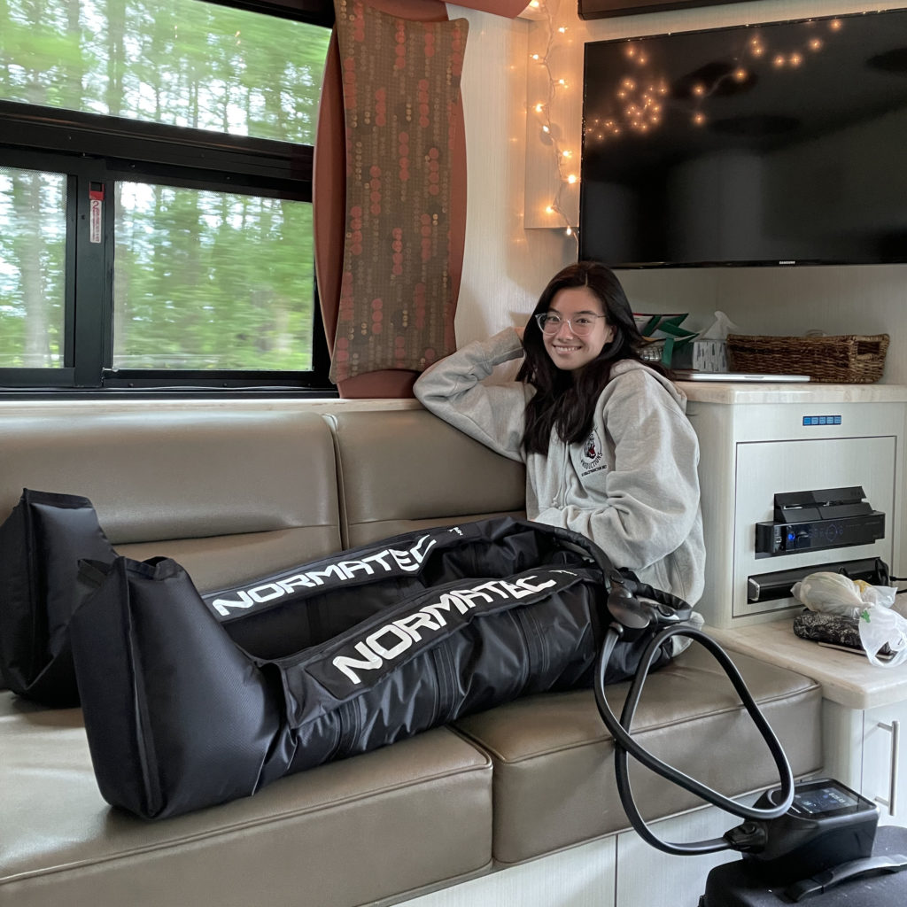 Chloe Misseldine sits on a leather banquet with her legs up on the cushions and smiles for the camera. She wears a sweatshirt, glasses and large, black compression boots on her legs.