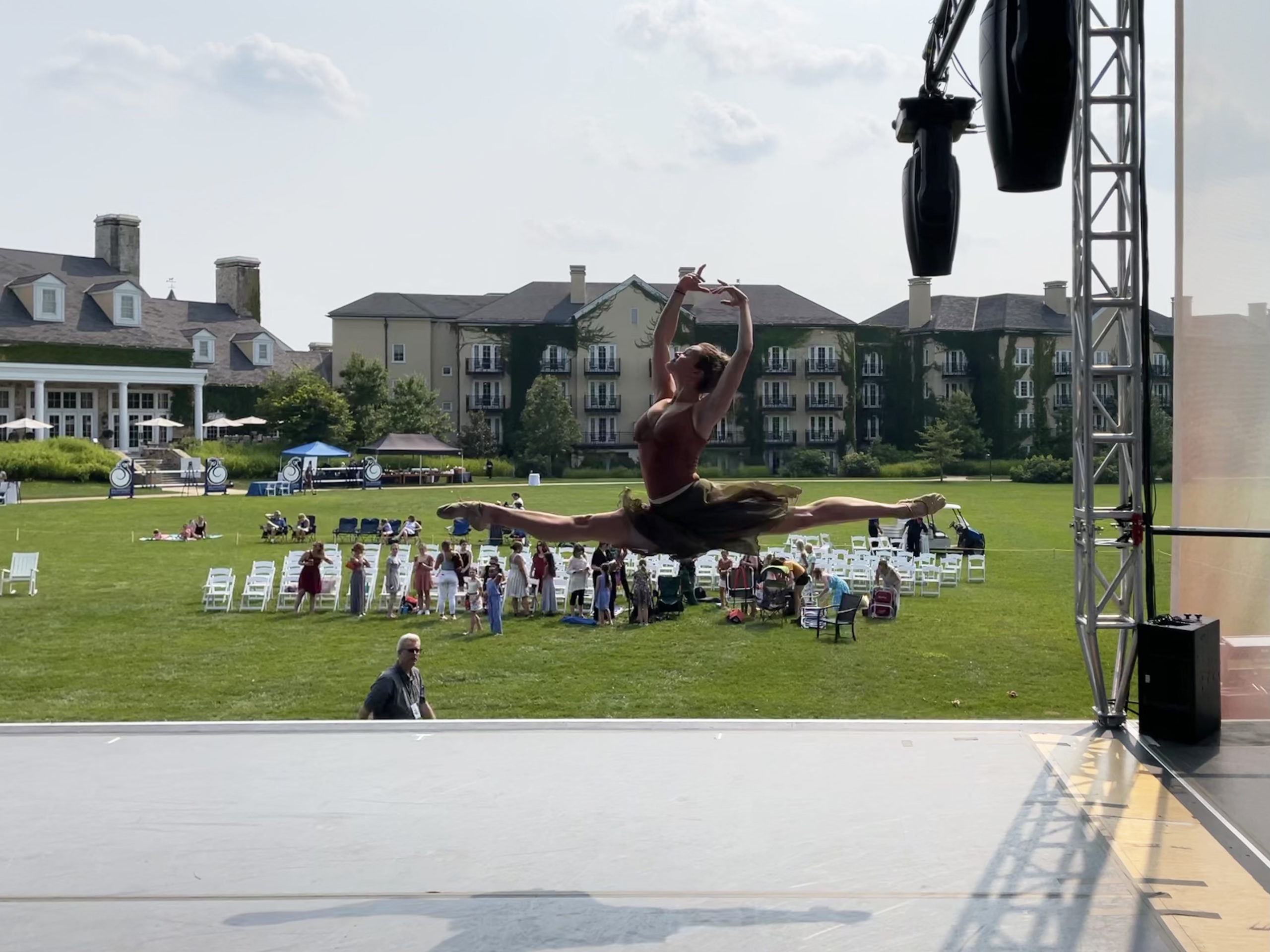 Abbey Marrison, wearing a brown leotard, black skirt and ballet slippers, does a saut de chat on an outdoor stage. A green lawn with white chairs and people milling about, as well as a large hotel complex, are in the background.