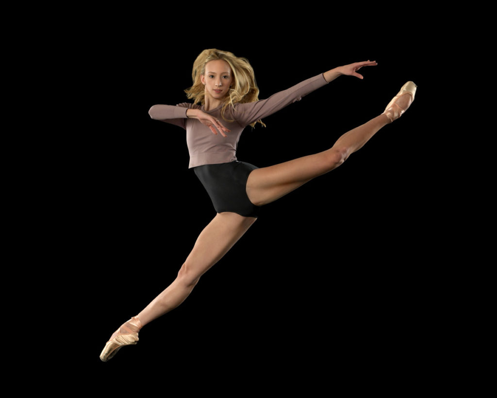 Ally Helman perfoms a sissone fermé with her back leg in a high attitude against a black backdrop. She croses her right arm across her chest and looks directly at the camera. Ally is wearing a black leotard with a mauve, long-sleeved crop top and pointe shoes.