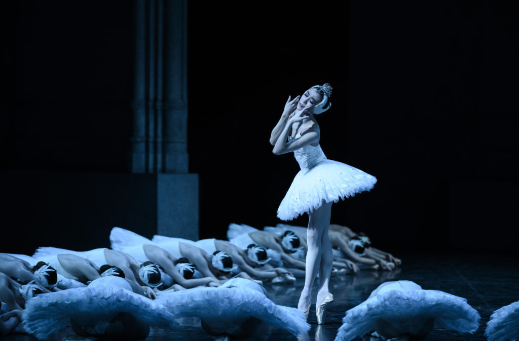 Sae Eun Park, dressed in costume as Odette/Odile in a white tutu and feathered tiara, bourreés in fifth and touches the sides of her face in anguish. A group of corps de ballet dancers poses on the ground around her.