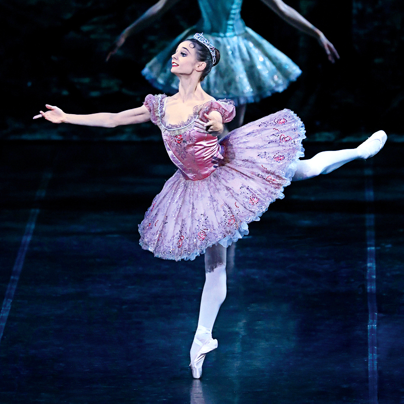 Maria Kochetkova wears a purple classical tutu, pink tights and pointe shoes and performs a piqué attitude effacé on her right leg with her arms opening to second. She looks out towards the corner and smiles regally.