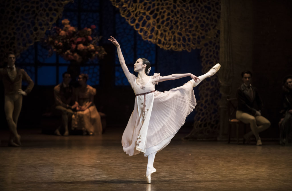 Wearing a pale pink dress, Sae Eun Park performs a first arabesque on pointe her right leg, a look of anguish on her face as she portrays Tatiana in Onegin. Behind her onstage, other dancers mill about the grand ballroom set.