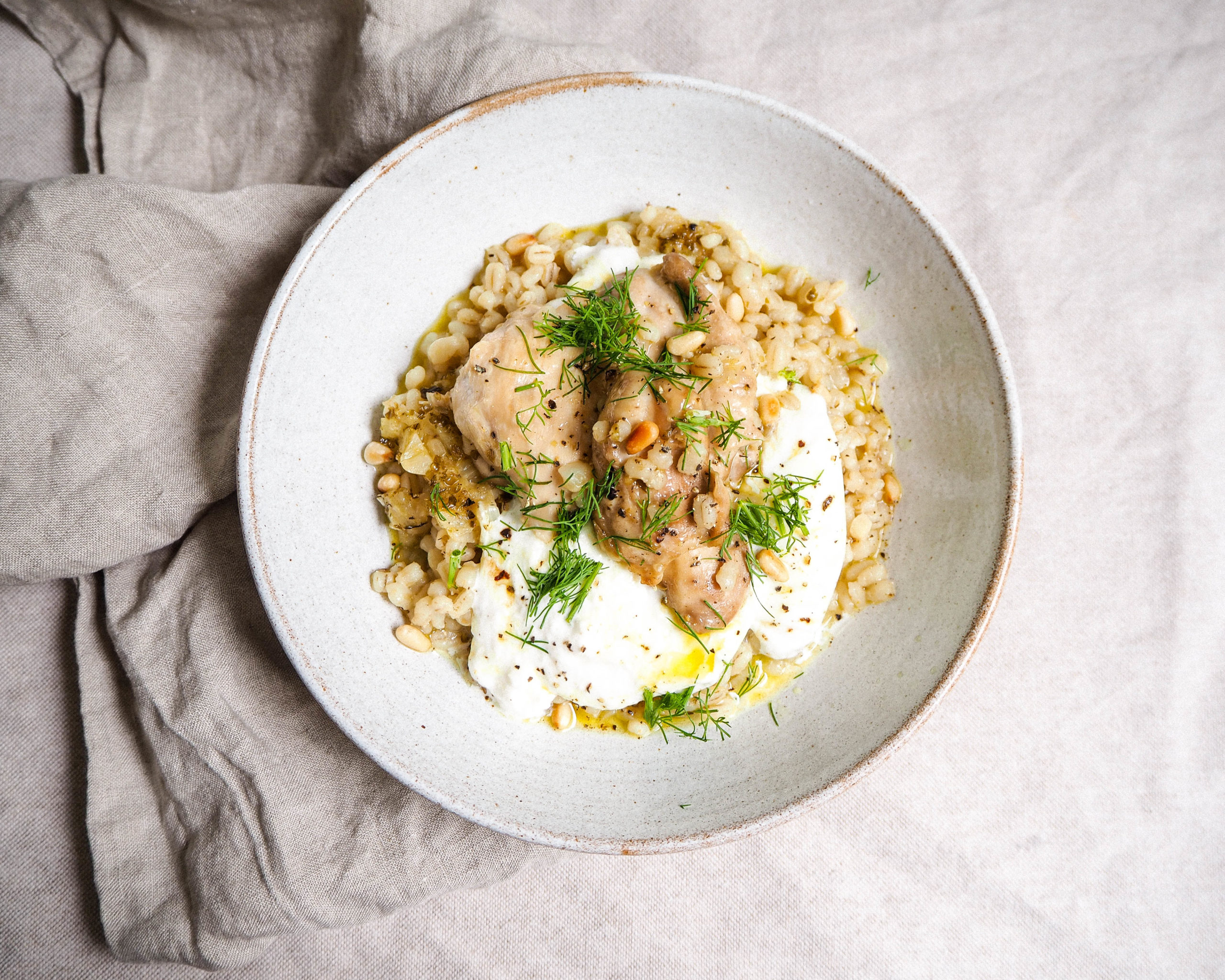 A white bowl is shown from above containing chicken over barley and buratta, with sprigs of dill on top.