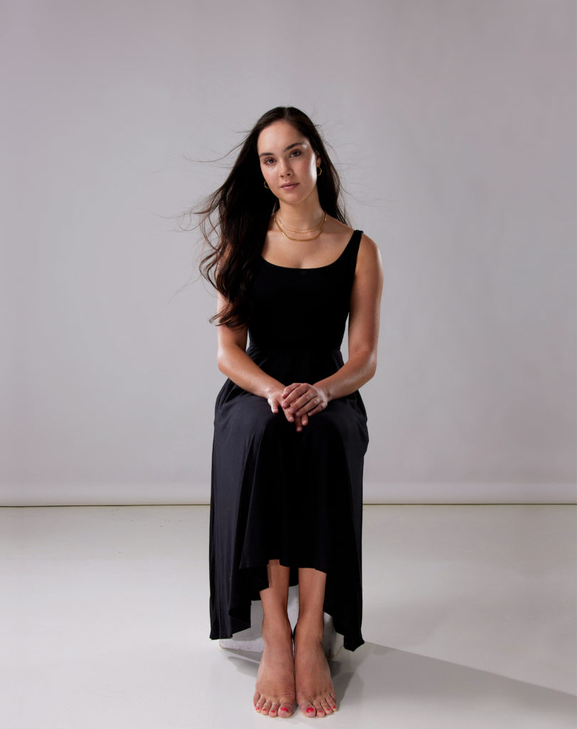 A barefoot Mira Nadon sits in a chair and tilts her head to the right, her hands resting gently on her knees. She wears a long, black jersey-knit dress and a simple gold necklace.