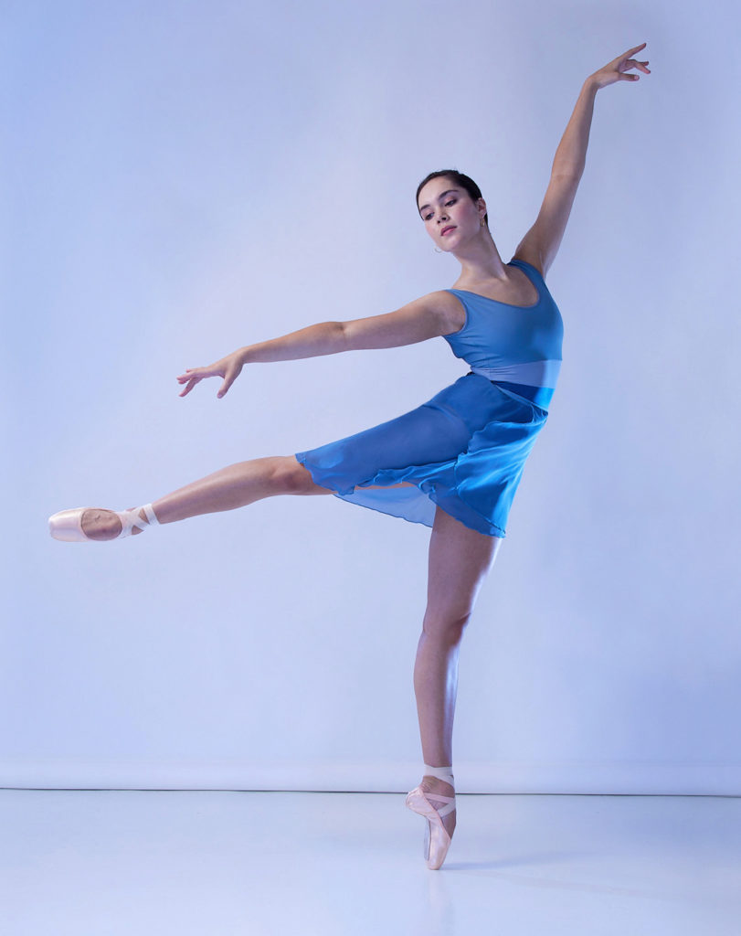 Mira Nadon poses in a low first arabesque on pointe and looks out over her right shoulder. She wears a blue leotard, short blue ballet skirt and pink pointe shoes in front of a cool blue backdrop.