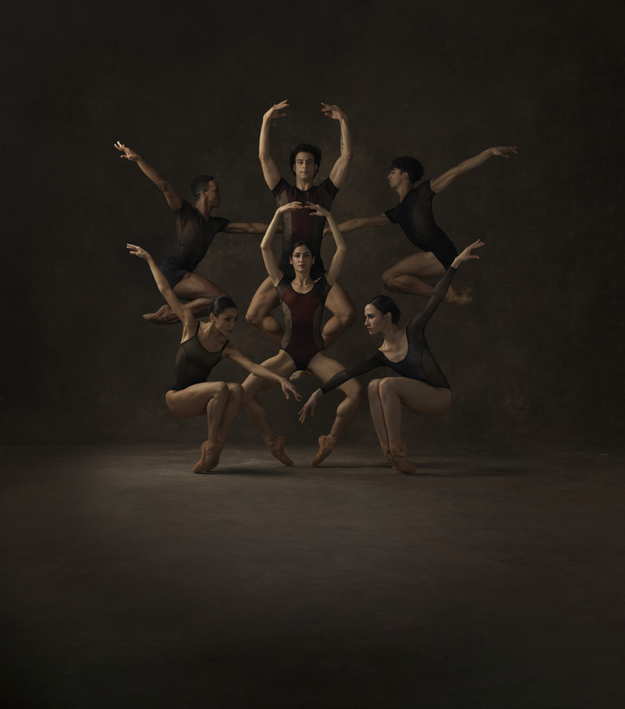 A group of male and female dancers in dark-colored leotards create a circular-shaped tableau. Some jump in the air with bent legs, while others create a similar shape pushing over their pointe shoes.