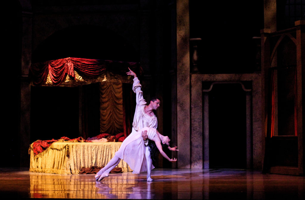 During a performance of Romeo and Juliet, Andres Castillo lunges to the left and holds his dance partner in his left arm as she swoons into a deep backbend. He is wearing a loose white tunic and white tights and she wears a pink nightgown, tights and pointe shoes. A bed is in the background onstage.